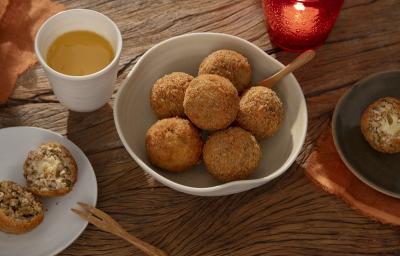 Foto em tons de laranja da receita de arancini de abobrinha servida em diversas porções em um bowl de cerâmica com uma colher de madeira sobre uma mesa de madeira. Ao lado há mais dois pratos com arancini e um copo de suco de laranja
