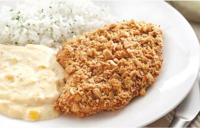Fotografia em tons de branco, amarelo e dourado, ao centro prato branco contendo creme de milho, arroz e frango empanado, ao lado uma faca e guardanapo cinza, sobre toalha listrada em tons ocre.