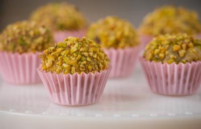 Fotografia de seis forminhas de cor rosa com brigadeiro de pistache enrolado no pistache triturado. Os brigadeiros estão em um apoio branco decorativo. Fotografia de seis forminhas de cor rosa com brigadeiro de pistache enrolado no pistache triturado. Os brigadeiros estão em um apoio branco decorativo.