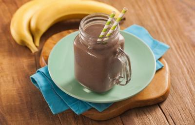 Fotografia em tons escuros, em uma bancada de madeira de cor marrom. Ao centro, uma tábua de madeira marrom contendo um pano azul. Em cima, há um prato verde e redondo contendo um copo com o milkshake. Ao fundo, há 2 bananas. Fotografia em tons escuros, em uma bancada de madeira de cor marrom. Ao centro, uma tábua de madeira marrom contendo um pano azul. Em cima, há um prato verde e redondo contendo um copo com o milkshake. Ao fundo, há 2 bananas.