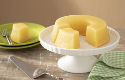 Foto em tons de amarelo da receita de bolo souza leão de pernambuco servida em uma base de porcelana alta circular com um prato de porcelana verde com um pedaço da receita. Ao lado há um paninho verde quadriculado e uma espátula prateada Foto em tons de amarelo da receita de bolo souza leão de pernambuco servida em uma base de porcelana alta circular com um prato de porcelana verde com um pedaço da receita. Ao lado há um paninho verde quadriculado e uma espátula prateada