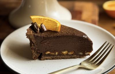 Fotografia de uma fatia de torta de chocolate e laranja com avelã em um prato pequeno raso branco. Ao lado da fatia tem um garfo apoiado no prato também. Fotografia de uma fatia de torta de chocolate e laranja com avelã em um prato pequeno raso branco. Ao lado da fatia tem um garfo apoiado no prato também.