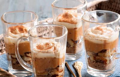 4 tacinhas transparentes com o doce banana com brigadeiro de caramelo e polvilhadas com canela. As 4 estão dentro de um cesto marrom com um pano azul. 4 tacinhas transparentes com o doce banana com brigadeiro de caramelo e polvilhadas com canela. As 4 estão dentro de um cesto marrom com um pano azul.