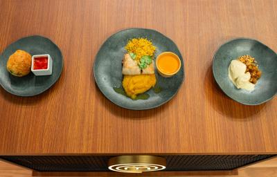 Fotografia em tons de marrom com três pratos cinzas ao centro. Dentro de um prato existe uma sobremesa de sorvete com amendoim, de outro existe um peixe e em outro uma entrada frita acompanhada de um molho de cor vermelha. Fotografia em tons de marrom com três pratos cinzas ao centro. Dentro de um prato existe uma sobremesa de sorvete com amendoim, de outro existe um peixe e em outro uma entrada frita acompanhada de um molho de cor vermelha.