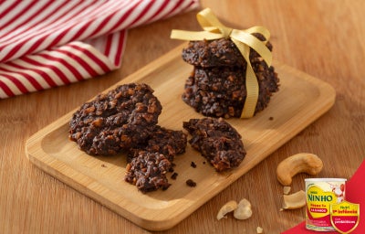 Cookies de cacau com castanhas sobre tábua de madeira, alguns quebrados, com castanhas ao redor e pano listrado ao fundo.