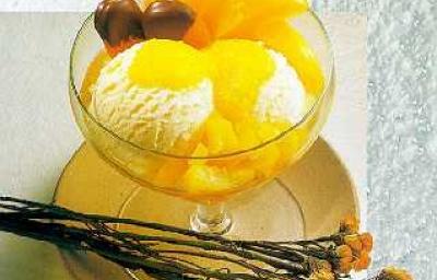 Fotografia em tons de amarelo em uma bancada de madeira, um pano bege, um prato redondo raso bege com uma taça de vidro com o sorvete de creme e pêssegos em calda dentro dela. Ao lado, flores amarelas e laranjas.