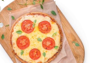 Fotografia em tons de amarelo e vermelho em uma mesa branca com uma tábua de madeira, um pano bege e uma pizza média de queijo com tomate em cima dela.