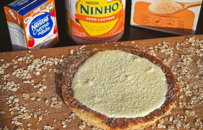 Fotografia em tons claros de uma tábua de madeira salpicada com farelo de aveia e tem uma panqueca de aveia com creme de ninho por cima e ninho salpicado. Atrás da tábua tem aveia e produtos zero lactose nestlé.