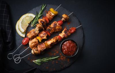 Fotografia vista de cima de três espetinhos, um do lado do outro, de carnes intercaladas com legumes. Os espetos estão sobre um apoio preto redondo, ao lado de duas fatias de limão siciliano, um molho vermelho em um recipiente menor preto e alecrim. Fotografia vista de cima de três espetinhos, um do lado do outro, de carnes intercaladas com legumes. Os espetos estão sobre um apoio preto redondo, ao lado de duas fatias de limão siciliano, um molho vermelho em um recipiente menor preto e alecrim.