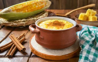 Fotografia de uma mini panela de barro apoiado em um apoio branco sobre uma mesa de madeira. Dentro da panelinha tem uma receita de canjica de milho verde com canela por cima. Ao redor tem paus de canela e um milho verde inteiro. Fotografia de uma mini panela de barro apoiado em um apoio branco sobre uma mesa de madeira. Dentro da panelinha tem uma receita de canjica de milho verde com canela por cima. Ao redor tem paus de canela e um milho verde inteiro.