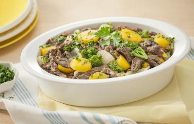 Fotografia em tons de amarelo em uma bancada de madeira clara com um pano amarelo e outro branco com listras azuis e um recipiente oval fundo com as tirinhas de carne com legumes dentro dele. Ao fundo, pratos rasos amarelos e um potinho com salsinha. Fotografia em tons de amarelo em uma bancada de madeira clara com um pano amarelo e outro branco com listras azuis e um recipiente oval fundo com as tirinhas de carne com legumes dentro dele. Ao fundo, pratos rasos amarelos e um potinho com salsinha.