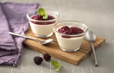 Fotografia em tons de branco e lilás, ao centro dois potinhos com creme branco e molho de framboesa e duas colheres sobre tábua de cozinha, ao lado guardanapo lilás, tudo sobre bancada em tom de lilás. Fotografia em tons de branco e lilás, ao centro dois potinhos com creme branco e molho de framboesa e duas colheres sobre tábua de cozinha, ao lado guardanapo lilás, tudo sobre bancada em tom de lilás.