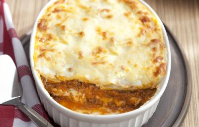 Fotografia em tons de branco e marrom, ao centro travessa branca com lasanha gratinada sobre tábua de cozinha, lado esquerdo guardanapo xadrez branco e vermelho, em bancada de madeira. Ao fundo, pedaço de lasanha em prato vermelho.