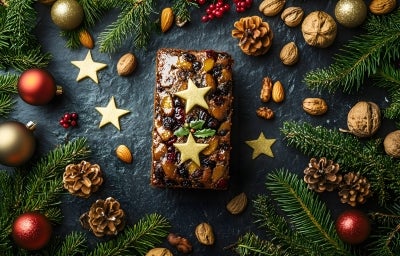 Fotografia de um bolo inglês decorado para as festas, com frutas secas, nozes e estrelas douradas, cercado por ramos de pinheiro e enfeites natalinos.