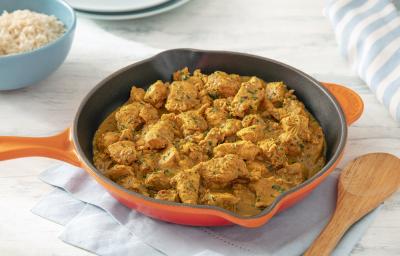 Foto da receita de frango ao curry sem lactose servida em uma frigideira grande laranja sobre uma mesa clara com um pano azul embaixo e uma colher de madeira ao lado Foto da receita de frango ao curry sem lactose servida em uma frigideira grande laranja sobre uma mesa clara com um pano azul embaixo e uma colher de madeira ao lado