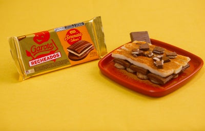 Sobremesa com banana, creme e chocolate recheado em prato vermelho, com embalagem Garoto ao lado.