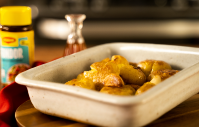 Foto da Receita de Batata Bolinha ao Murro. Observa-se um recipiente refratário de cerâmica com as batatas em cima de uma tábua de madeira. Ao fundo, um pote de Maggi Fondor.