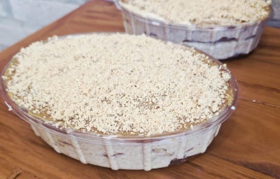 Tortas em embalagens plásticas, cobertas com farofa crocante e creme claro, sobre mesa de madeira.