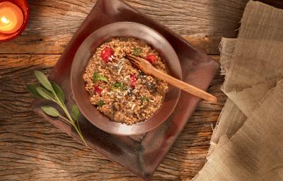 Foto em tons de marrom da receita de risoto ao funghi com aveia corte rústico servida em um prato fundo marrom sobre uma base retangular de mesma cor em cima de uma mesa de madeira