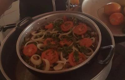 Fotografia de uma panela grande com moqueca com tomates, pimentões e cebolas em rodelas por cima. A panela está dentro de uma outra panela maior. Fotografia de uma panela grande com moqueca com tomates, pimentões e cebolas em rodelas por cima. A panela está dentro de uma outra panela maior.