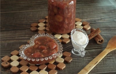 Fotografia em tons escuros de uma mesa de madeira escura. Sobre a mesa tem dois apoios quadriculados de madeira, apoiado em um deles tem um pote de vidro com doce de banana. No outro apoio tem o doce num pote de vidro pequeno em formato de coração.