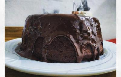 Fotografia em tons de preto em uma mesa de madeira, um pano vermelho, um prato branco raso redondo com o bolo de chocolate com cobertura de chocolate em cima. Fotografia em tons de preto em uma mesa de madeira, um pano vermelho, um prato branco raso redondo com o bolo de chocolate com cobertura de chocolate em cima.