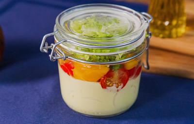 Fotografia em tons de azul com um pote de vidro ao centro. Dentro do pote existe uma salada feita com manga, alface, tomate cereja e molho de mostarda com iogurte