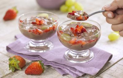 Fotografia em tons de branco e roxo de uma bancada de madeira branca com um paninho roxo, sobre ele duas taças de frutas com creme e uma colher retirando um pedaço. Ao fundo uvas e morangos. A frente dois morangos. Fotografia em tons de branco e roxo de uma bancada de madeira branca com um paninho roxo, sobre ele duas taças de frutas com creme e uma colher retirando um pedaço. Ao fundo uvas e morangos. A frente dois morangos.