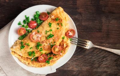 fotografia em tons de marrom, branco, amarelo e vermelho de uma bancada marrom vista de cima. Contém um prato redondo branco com uma omelete, e por cima pedaços de tomates e salsas fotografia em tons de marrom, branco, amarelo e vermelho de uma bancada marrom vista de cima. Contém um prato redondo branco com uma omelete, e por cima pedaços de tomates e salsas