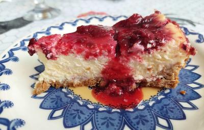 Foto em tons de vermelho da receita de cheesecake de frutas vermelhas ao estilo nova york servida em uma fatia sobre um prato decorado de porcelana azul e branco