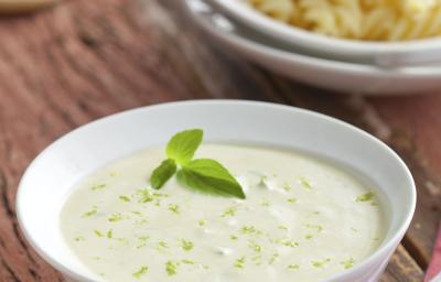 Fotografia com tons de branco e marrom, com ramequim branco com molho, ao fundo prato branco com macarrão parafuso e pedaço de queijo com ralador sobre tábua branca, ao lado guardanapo listrado, tudo sobre bancada de madeira rústica.,