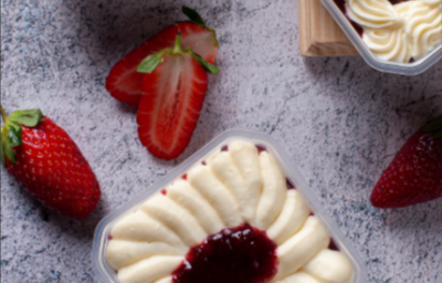 Imagem mostra, de cima, dois recipientes quadrados transparentes com um creme branco e um creme vermelho no topo. Estão dispostos em uma bancada de granito cinza, com uma tábua de madeira com morangos inteiros, cortados jogados.