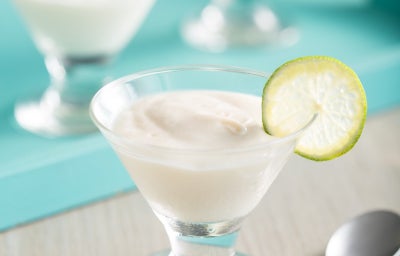 Foto de uma taça de vidro com sobremesa cremosa de limão, de cor clara e textura lisa. Uma fatia fina de limão está apoiada na borda. Ao fundo, há outras taças semelhantes sobre bandeja azul. A cena inclui colher prateada e mesa clara