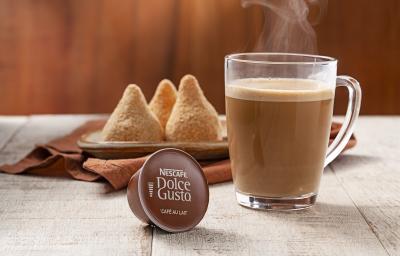 Fotografia em tons de marrom, cinza e branco de uma bancada cinza vista de frente, contém uma capsula de café ao lado uma xicara transparente com café. Ao fundo um pano marrom com um prato marrom e 3 coxinhas por cima