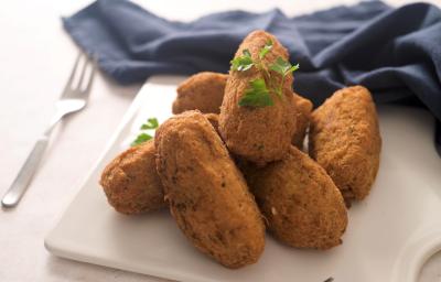 Fotografia de vários bolinhos de bacalhau fritos sobre um apoio branco, ao lado de um pano azul de tom escuro e um garfo.