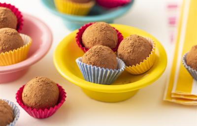 Fotografia em tons de marrom, em uma bancada de madeira branca. Ao centro, há um recipiente redondo amarelo contendo 3 bananas trufadas dentro de forminhas. Ao lado, há algumas bananas trufas espalhadas e um pano listrado amarelo. Fotografia em tons de marrom, em uma bancada de madeira branca. Ao centro, há um recipiente redondo amarelo contendo 3 bananas trufadas dentro de forminhas. Ao lado, há algumas bananas trufas espalhadas e um pano listrado amarelo.
