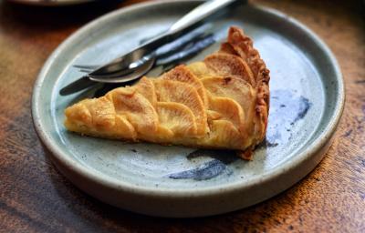 Fotografia de uma fatia de torta de maçã caramelada e recheada com pedaços de maçã. Ao lado da torta tem talheres que também estão por cima de um prato raso de cerâmica em tom de azul, e ele está sobre uma mesa de madeira.