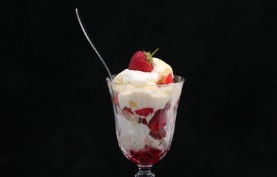 Fotografia de uma taça de vidro com merengue e morangos, de forma intercalada. E dentro do recipiente, uma colher de sobremesa apoiada, sobre um fundo preto. Fotografia de uma taça de vidro com merengue e morangos, de forma intercalada. E dentro do recipiente, uma colher de sobremesa apoiada, sobre um fundo preto.