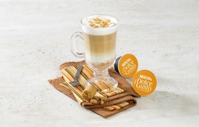 fotografia em tons de branco e marrom de uma bancada branca vista de cima. Contém um xicara transparente com a bebida de café e paçoca dentro, abaixo um pano em tons de marro, bege e laranja e duas capsula de Nescafé Dolce Gusto fotografia em tons de branco e marrom de uma bancada branca vista de cima. Contém um xicara transparente com a bebida de café e paçoca dentro, abaixo um pano em tons de marro, bege e laranja e duas capsula de Nescafé Dolce Gusto