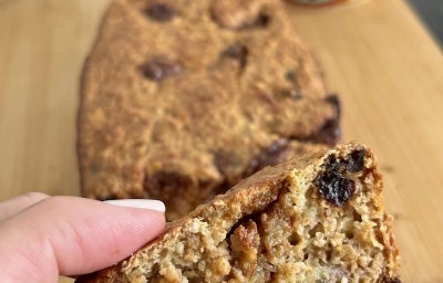 Fotografia de uma mão feminina, com unhas pintadas de branco, segurando um pedaço do bolo de banana, aveia e ameixa. Fotografia de uma mão feminina, com unhas pintadas de branco, segurando um pedaço do bolo de banana, aveia e ameixa.