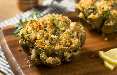Fotografia de duas alcachofras gratinadas com farofa e salsinha por cima, sobre uma tábua de madeira em tom escuro que está apoiada em um pano branco com listras pretas. Ao lado, fatias de limão siciliano.