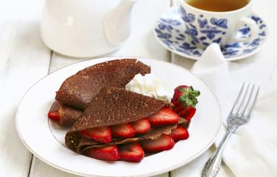 Fotografia de panquecas de chocolate com morango, e um pouco de chantilly ao lado, dentro de um prato branco raso. Ao redor , um garfo sobre uma toalha branca, um bule branco e uma xícara de chá em cima do prato pequeno, ambos são azuis e brancos. Fotografia de panquecas de chocolate com morango, e um pouco de chantilly ao lado, dentro de um prato branco raso. Ao redor , um garfo sobre uma toalha branca, um bule branco e uma xícara de chá em cima do prato pequeno, ambos são azuis e brancos.