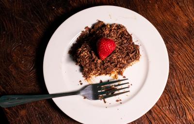 Fotografia vista de cima de um pedaço de pavê com mousse e chocotrio. Por cima tem ganache, raspas de chocolate e morango. O pavê está sobre um prato branco de vidro com um garfo.