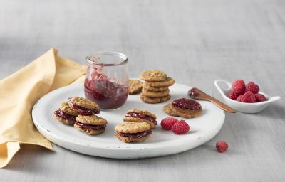 fotografia em tons de cinza, bege e vermelho de uma bancada cinza vista de cima. Ao centro um prato branco redondo com um pote transparente com geleia e em volta vários Whoopies recheado com a geleia. Do lado esquerdo um pano bege. fotografia em tons de cinza, bege e vermelho de uma bancada cinza vista de cima. Ao centro um prato branco redondo com um pote transparente com geleia e em volta vários Whoopies recheado com a geleia. Do lado esquerdo um pano bege.