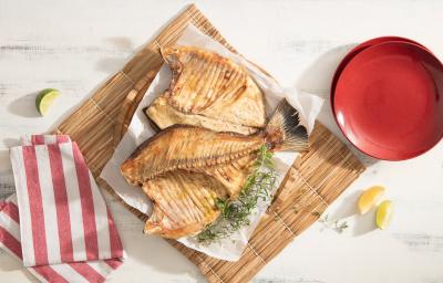 Fotografia em tons de vermelho em uma mesa de madeira branca com um jogo americano de palha ao centro, com um papel manteiga e a costela de tambaqui assada em cima dele. Ao lado, um pano branco com listras vermelhas. Fotografia em tons de vermelho em uma mesa de madeira branca com um jogo americano de palha ao centro, com um papel manteiga e a costela de tambaqui assada em cima dele. Ao lado, um pano branco com listras vermelhas.