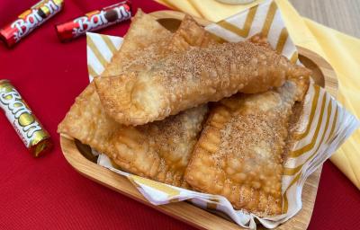 Fotografia em tons de vermelho com um recipiente com vários pastéis recheados com chocolate Baton ao centro. Os pastéis estão polvilhados com açúcar e canela. Ao fundo existe embalagens de chocolate Baton. Fotografia em tons de vermelho com um recipiente com vários pastéis recheados com chocolate Baton ao centro. Os pastéis estão polvilhados com açúcar e canela. Ao fundo existe embalagens de chocolate Baton.