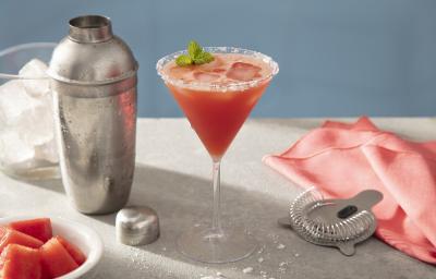 Foto em tons de vermelho da receita de margarita de melancia servida em uma taça de vidro sobre uma mesa de mármore onde estão uma coqueteleira cromada, além de um pano vermelho, uma mola para mistura, um copo com gelo e pedaços de melancia em cubos. Foto em tons de vermelho da receita de margarita de melancia servida em uma taça de vidro sobre uma mesa de mármore onde estão uma coqueteleira cromada, além de um pano vermelho, uma mola para mistura, um copo com gelo e pedaços de melancia em cubos.