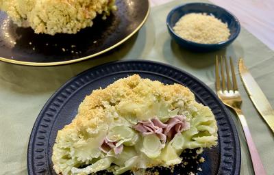 Foto em tons de amarelo claro da receita de couve-flor recheada servida em um prato de porcelana azul escuro com talheres prateados ao lado e um recipiente azul com queijo parmesão ralado. Ao fundo há um prato preto com outra porção da couve-flor