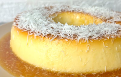 Fotografia em tons claros com um pudim de coco ao centro. O pudim ter cor caramelada e é coberto com coco ralado por cima.