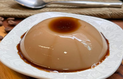 Imagem mostra um pudim redondo e pequeno, coberto de caramelo, em tons castanhos e marrons sobre um prato branco. O prato está apoiado em cima de uma madeira, com grãos de café espalhados, um guardanapo de pano bege e uma colher.